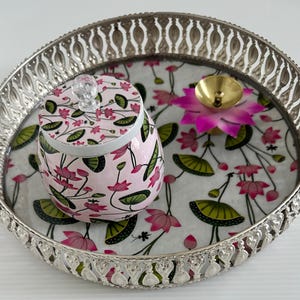 May include: A silver-colored, round decorative tray with a floral design. The tray holds a pink jar with a floral pattern, a small gold-colored bowl, and a pink lotus flower. The tray's rim has a detailed, openwork design.