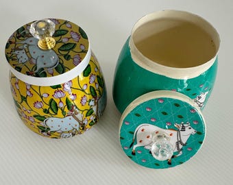 May include: Two decorative jars with lids. One jar is yellow with a floral and cow design, and the other is green with a cow design on the lid. Both lids have crystal knobs.