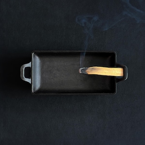 May include: A rectangular, black cast iron incense burner with two handles. A lit palo santo stick is placed inside, emitting a wisp of smoke against a dark background. The burner is designed for burning incense.