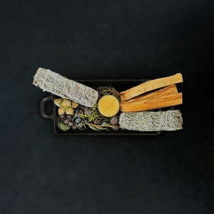 May include: A rectangular black tray containing two white sage bundles, two light brown palo santo sticks, a small yellow candle, and a collection of dried green foliage, small dark berries, and a single light-colored dried flower blossom.