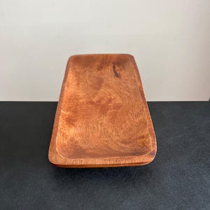May include: A rectangular wooden tray with rounded corners. The tray is made of a reddish-brown wood, with visible grain patterns. The tray is empty and sits on a dark surface, with a white background.