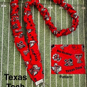May include: Red Texas Tech lanyard with a repeating pattern of the Texas Tech logo and the words "Texas Tech" and "Red Raiders". The lanyard has a silver-colored metal clasp and key ring. The background is a green field.