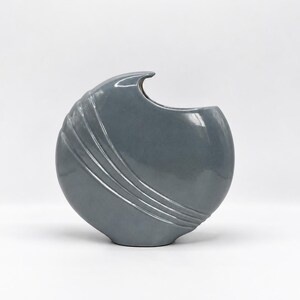 May include: A circular, slate-gray ceramic vase with a glossy finish. The vase features a crescent-shaped opening at the top and three distinct, raised diagonal ridges that curve along the left side, creating a textured, wave-like appearance.