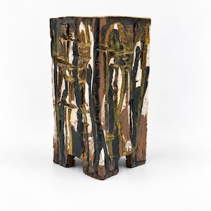 May include: A rectangular ceramic vessel with a textured, uneven surface featuring a glaze of brown, black, white, and mustard yellow. The object has four short, blocky feet at the base and vertical, drip-like patterns across its four sides.