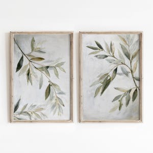 May include: Two rectangular wall art prints featuring painted olive branches with muted green and grey leaves against a textured light grey background. Each print is encased in a thin, light-toned wooden frame, displaying botanical art.