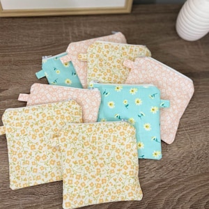 May include: A collection of small, zippered pouches in various floral patterns. The pouches feature a mix of colors, including yellow, pink, and teal, with delicate flower designs. Each pouch has a small loop for easy carrying.