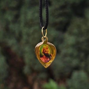 Puede incluir: Un colgante en forma de lágrima que presenta el retrato de una figura barbuda con turbante, encerrado en un marco de tono dorado. El colgante cuelga de un cordón negro, con la imagen central sobre un fondo verde dentro del borde dorado.