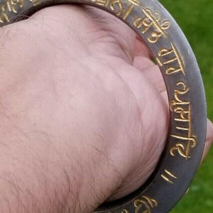 Peut inclure: Un bracelet circulaire en métal gris foncé orné d'une inscription en écriture gurmukhi gravée et remplie d'une incrustation dorée. Le bracelet présente une finition mate lisse et une épaisseur constante sur toute sa bande arrondie.