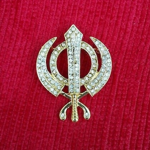 Puede incluir: Un colgante Sikh Khanda de color dorado con adornos claros en forma de cristal. El colgante presenta una espada de doble filo, dos Kirpans curvados y un Chakkar circular. El colgante está sobre un fondo rojo.
