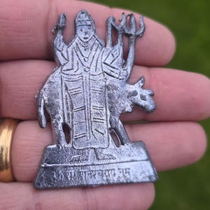 Puede incluir: Figura religiosa de metal plateado que representa una deidad montada en un animal, sosteniendo un tridente y otros objetos. La figura tiene detalles intrincados y está montada sobre una base rectangular con texto. El fondo es verde borroso.