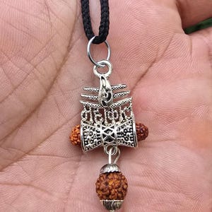 May include: Silver pendant necklace with a black cord. The pendant has detailed patterns and two small, round, brown beads. A larger, textured brown bead hangs below the pendant. The necklace is a spiritual item.