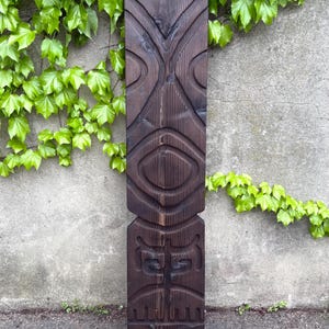 May include: A dark brown, carved wooden tiki statue with a weathered appearance. The statue features a stylized face with prominent eyes and a mouth, and is approximately 60 inches tall. The background includes green ivy on a gray wall.