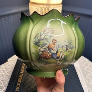 May include: Green ceramic planter with a scalloped rim and a pastoral scene. The planter features a glossy finish and a decorative illustration of people and sheep. The rim has a gold trim.