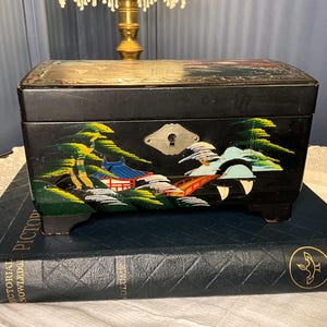May include: A rectangular black lacquered jewelry box featuring a hand-painted landscape with a red bridge, green trees, and a building. The box has a silver-toned metal keyhole plate on the front and sits on four small feet.