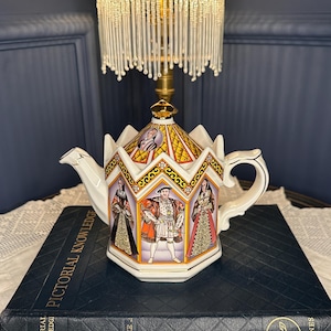 May include: A hexagonal ceramic teapot featuring a gold-trimmed lid and panels depicting historical figures in royal attire. The teapot has a white handle and spout, with intricate orange, yellow, and black geometric patterns bordering each portrait.