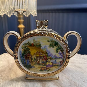 May include: Cream ceramic sugar bowl with gold detailing and handles. The oval centrepiece depicts a cottage scene with a thatched roof, flowers, and a figure. The lid has a gold bow-shaped handle.