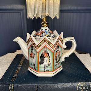 May include: A hexagonal ceramic teapot featuring colourful illustrations of historical military figures in uniform on each panel. The lid is topped with a gold-coloured knob, and the body is decorated with geometric patterns in red, blue, and gold.