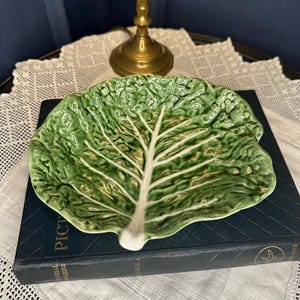 May include: A glossy ceramic dish shaped like a green cabbage leaf with a prominent white central vein and textured, ruffled edges. The dish features a vibrant green glaze with lighter and darker tones, mimicking the natural appearance of a leaf.