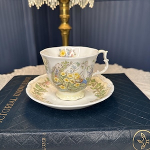 May include: A white porcelain teacup and saucer set featuring a delicate floral pattern with yellow daffodils and green foliage. The teacup has a scalloped rim and a decorative handle, while the saucer displays a matching botanical design.