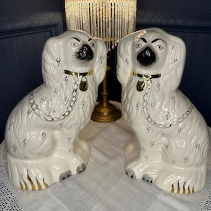 Peut inclure: Une paire de figurines de chiens épagneuls en céramique blanche avec des détails faciaux peints en noir, des colliers dorés avec des médailles suspendues et des motifs de chaînes dorées peints sur le corps. La base est ornée de rayures dorées.