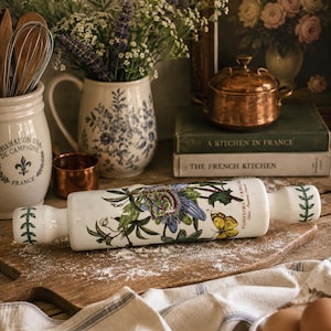 Può includere: Un mattarello in ceramica bianca con un motivo floreale raffigurante un fiore della passione e una farfalla gialla. Il mattarello è appoggiato su un tagliere di legno cosparso di farina.