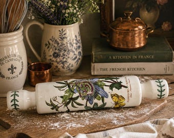 May include: A white ceramic rolling pin with a floral design featuring a passion flower and a yellow butterfly. The rolling pin rests on a wooden cutting board dusted with flour, alongside a linen towel.