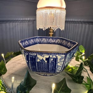 May include: A blue and white ceramic bowl featuring a cathedral design. The hexagonal bowl has a gold rim. A vintage lamp with a beaded fringe is positioned above. The bowl is decorated with greenery and fairy lights.