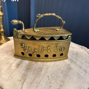 Puede incluir: Antigua plancha de latón con asa curvada y diseño floral decorativo. La plancha tiene una tapa con bisagras y una serie de pequeños agujeros en la parte delantera. El asa y la tapa están adornadas con detalles intrincados.