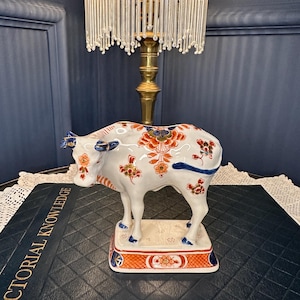 Può includere: Una statuetta di mucca in ceramica bianca con motivi floreali dipinti a mano in arancione, blu e oro. La mucca poggia su una base rettangolare con un bordo arancione a tratteggio incrociato e accenti blu. La superficie è lucida e dettagliata.