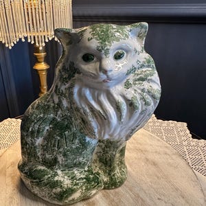 May include: A ceramic cat ornament with a white base and green mottled pattern. The cat has green eyes and a detailed, textured fur design. The figurine is sitting on a wooden surface, with a lamp in the background.