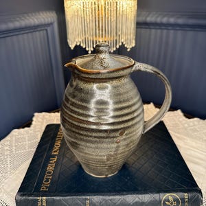 May include: A stoneware pitcher with a lid, featuring a mottled grey and brown glaze with horizontal ribbed textures. The pitcher has a curved handle and a small spout, resting on a dark blue book titled Pictorial Knowledge by Newnes.