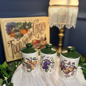 May include: Three ceramic canisters with green lids and fruit illustrations. The canisters feature images of peaches, plums, and apples. The canisters are arranged on a white wooden tray. A book titled "Food From Your Garden" is in the background.