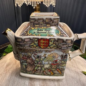 Può includere: Una teiera in ceramica bianca con coperchio a forma di castello, con un'illustrazione colorata di Robin Hood nella foresta di Sherwood. La teiera ha dettagli dorati e una scena dettagliata di personaggi con archi e frecce.