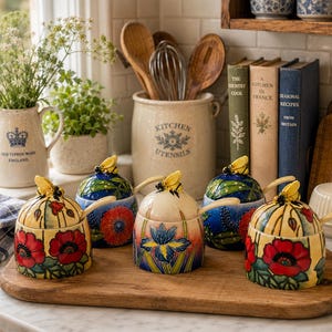 May include: Five ceramic honey pots with lids, each featuring a small bee figurine on top and a matching spoon. The pots display colourful hand-painted floral designs including red poppies and blue irises on cream and blue backgrounds.