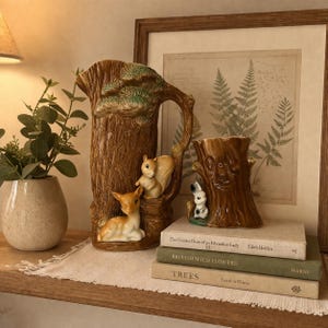 Può includere: Un set di brocca e vaso in ceramica marrone, entrambi a forma di tronchi d'albero, con figurine di animali del bosco. La brocca presenta un cervo e uno scoiattolo, mentre il vaso ha un coniglio. Libri e una stampa di felci incorniciata completano l'esposizione.