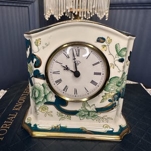 Puede incluir: Un reloj de chimenea de cerámica color crema con una esfera redonda y números romanos. El reloj está decorado con motivos florales verdes y ribetes dorados. El reloj mide aproximadamente 18 cm de alto.
