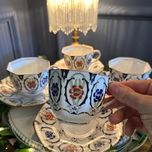 Può includere: Un set di tazze e piattini vintage. Le tazze e i piattini in porcellana bianca presentano un motivo floreale rosso, blu e oro. Le tazze hanno una forma ottagonale. Il set è esposto su un vassoio decorativo.