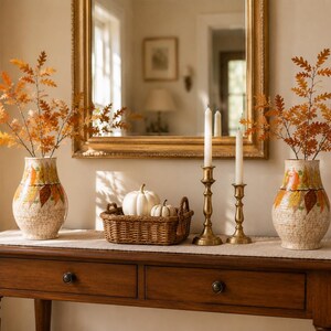 May include: Two ceramic vases with a textured cream finish and a painted autumn leaf pattern, each holding orange oak branches. Between them sits a woven brown basket containing two white pumpkins and two brass candlesticks with white candles.