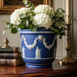 May include: A deep blue ceramic planter featuring white relief ornamentation of lion heads, floral swags, and classical figures. The vessel is filled with a large arrangement of white hydrangea blossoms and green eucalyptus foliage.
