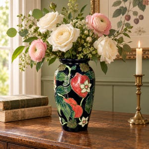 May include: A dark blue ceramic vase featuring a raised, hand-painted design of red strawberries, green leaves, and white blossoms. The vase is filled with a bouquet of white and light pink roses and ranunculus flowers.