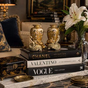 May include: Two ornate gold vases with serpent-like handles sit atop a stack of books with the titles "Chanel", "Valentino", and "Vogue". A black lacquered box with gold accents and a small brass dish are in the foreground.