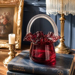 May include: A deep red glass basket featuring a ruffled, crimped rim and a clear, textured glass handle. The basket has a wide, rounded base that tapers slightly towards the top, showcasing a translucent, vibrant crimson colour throughout the glass.