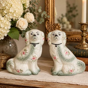 May include: A pair of ceramic dog figurines featuring a white base with hand-painted pink floral patterns, green accents, and a black collar. The dogs are depicted in a seated position with detailed fur textures and expressive facial features.