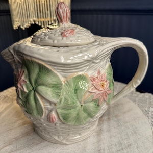 May include: A white ceramic teapot with a basket weave design, featuring pink water lilies and green lily pads. The teapot has a curved handle and a lid with a pink bud-shaped finial. The teapot is a decorative item.