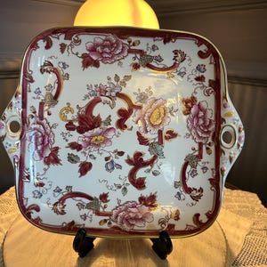 Peut inclure: Plateau de service carré à motifs floraux. Le plateau présente un fond blanc orné de motifs floraux roses, rouges et dorés. Il possède une bordure bordeaux et deux poignées avec des ouvertures circulaires. Le plateau est présenté sur un support noir.