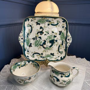 Pode incluir: Uma bandeja de servir quadrada de cerâmica com alças, uma tigela a condizer e uma leiteira. Cada peça apresenta uma base de cor creme com um padrão floral detalhado em verde e azul-petróleo escuro e bordas douradas no contorno.