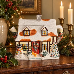 May include: A ceramic teapot shaped like a cottage with a snow-covered roof, a red front door with a wreath, a snowman, and a decorated Christmas tree. The base features the text CHRISTMAS COTTAGE. It has a chimney lid and a handle.