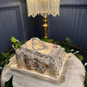 May include: A vintage, rectangular covered dish with a floral design. The dish is white with gold trim and a handle on top. The floral design includes pink, blue, and yellow flowers. The dish sits on a matching rectangular plate.