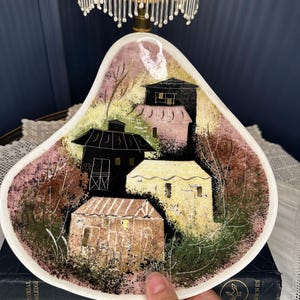 May include: A teardrop-shaped ceramic dish featuring a painted scene of three houses in black, cream, and brown tones. The background includes textured pink, green, and yellow brushstrokes, with thin black lines depicting trees and foliage surrounding the structures.