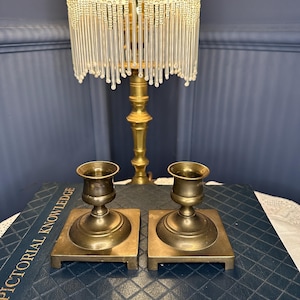 Op de afbeelding: Twee antieke messing kandelaars met vierkante voet. De kandelaars staan op een donkerblauw boek met de woorden "Pictorial Knowledge" zichtbaar. Een vintage messing lamp met een kralenfranje staat op de achtergrond.